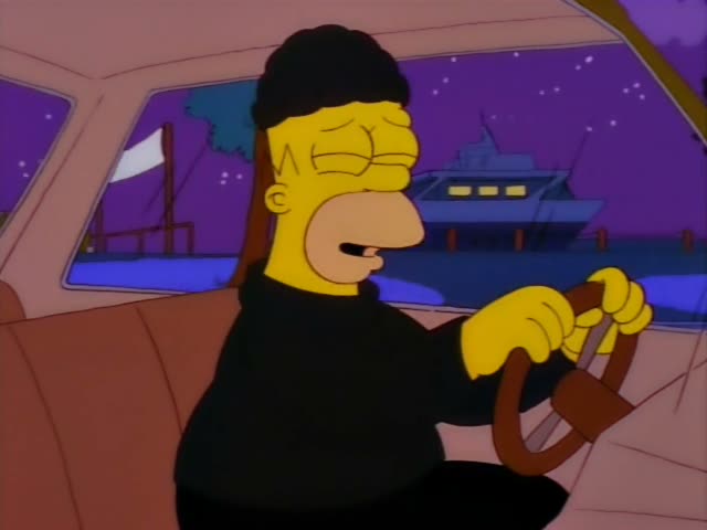 A heavyset yellow-skinned man in a black knit cap and black sweater grips the steering wheel of a tan car at night…