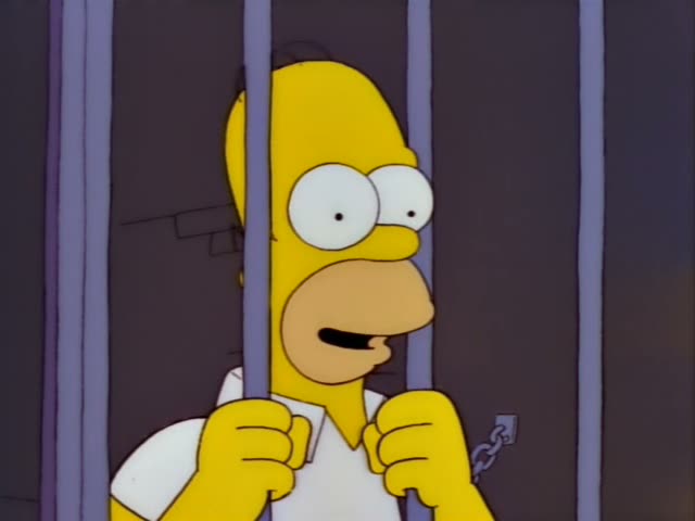 Homer Simpson stands behind jail cell bars, gripping them with both hands and a chain dangling from his wrist, first…