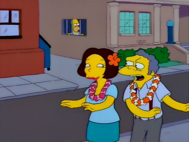 Moe and a brown-haired woman with an orange hibiscus in her hair, both wearing Hawaiian leis, stand on a sidewalk in front…