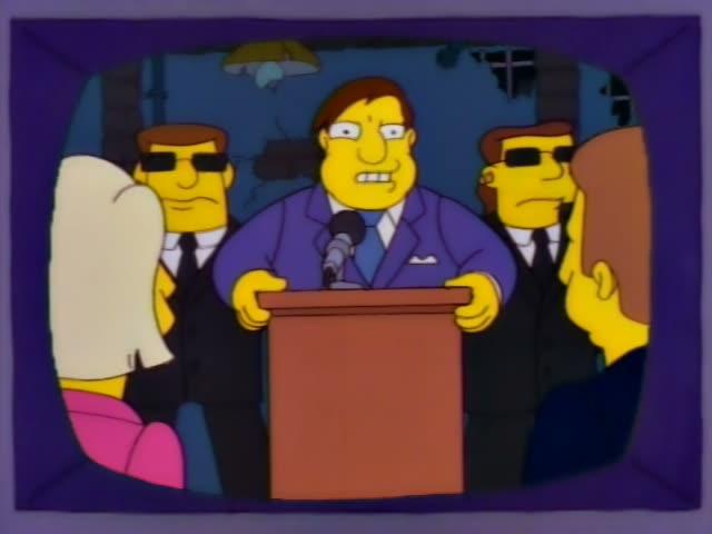 A heavy, bald-topped man with brown hair in a purple suit and blue tie stands behind a wooden podium with a microphone…