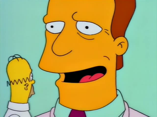 Troy McClure appears on a large screen in a dark hall while Marge stands at a rail beside Homer, Lisa and Bart