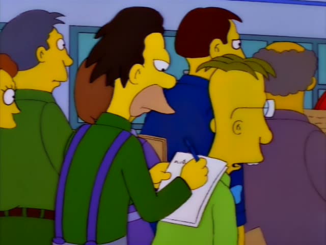 A crowd stands in a hallway lined with lockers, with a bearded man in a green sweater and purple suspenders writing…