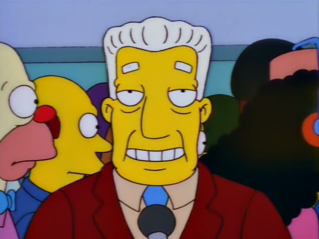 A white-haired, red-jacketed older man with a blue tie stands at a microphone in the foreground, with a bald heavy man…
