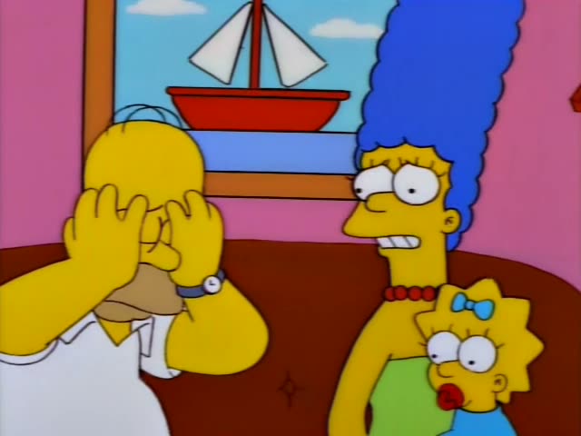 Homer Simpson sits on a brown couch in the living room with Marge, who has a tall blue beehive and a red bead necklace…