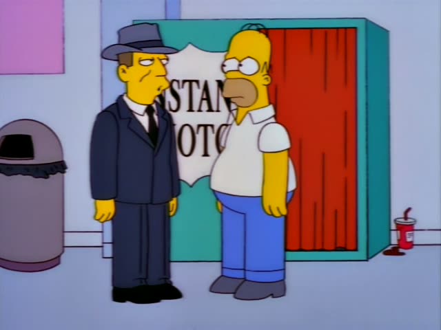 Homer stands beside a man in a dark suit and fedora in front of an Instant Photos booth with a red curtain and a red cup…