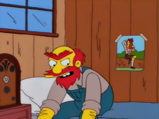 A red-bearded, red-haired man in green overalls and a grey shirt sits on a bed in a wood-panelled room with a blue-paned…
