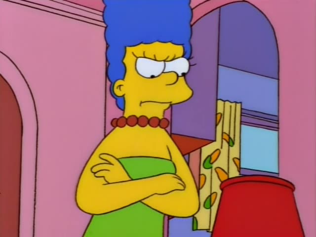 Marge Simpson stands in a green dress with her arms crossed in front of a pink wall, an arched doorway, and a red lamp…