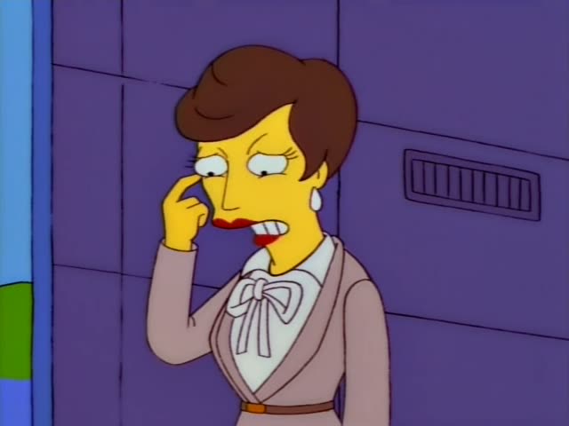 A short-haired brown-haired woman in a beige suit with a white bow blouse and red lipstick stands against a purple wall…