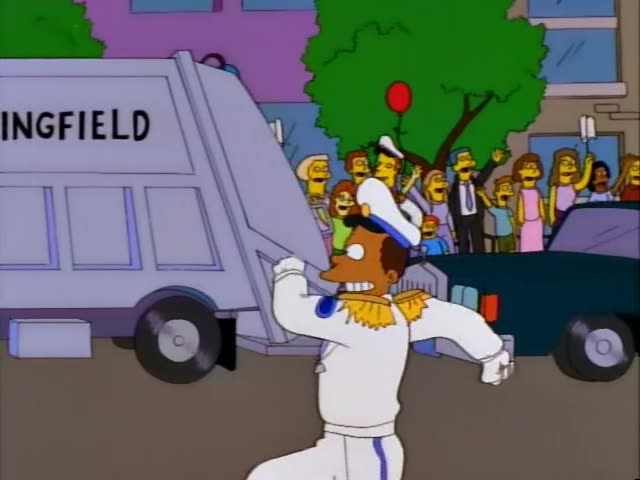 Homer Simpson rides atop a Springfield garbage truck through a parade on a city street, with the crowd lined…
