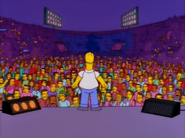 Homer Simpson stands on a stage before a huge crowd with his arms spread, then turns to face a thin man with slicked-back…
