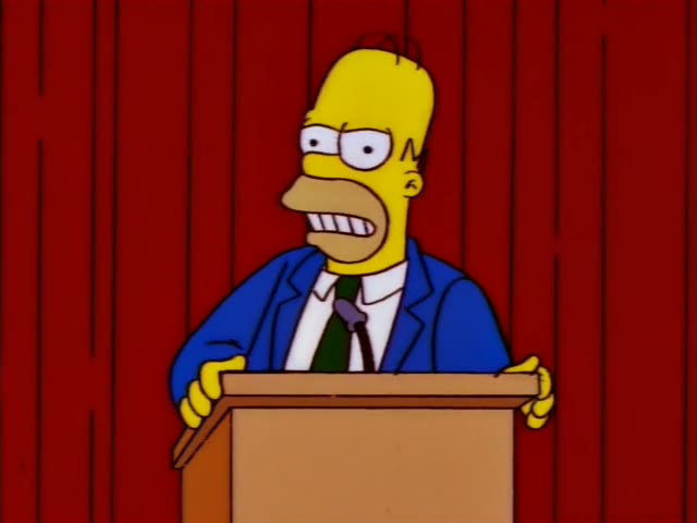Homer Simpson stands at a wooden podium before red curtains, wearing a blue jacket and green tie, grimacing and gesturing…
