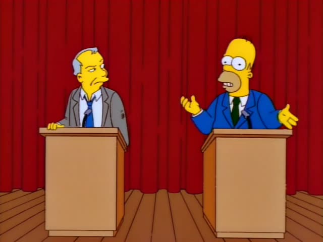 Homer Simpson stands at the right of two wooden podiums on a stage with a red curtain, facing a grey-haired man in a grey…