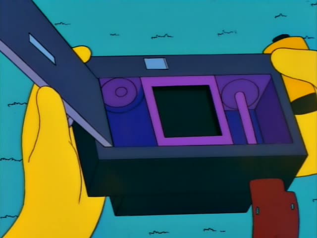 Marge Simpson holds a black handheld device with a strap in a pink room, first looking off to the side and then raising it…
