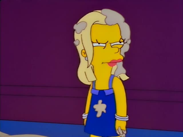 A blonde woman with a grey-streaked beehive-like hairdo and a blue dress with a tan patch, wearing bracelets and blue heels…
