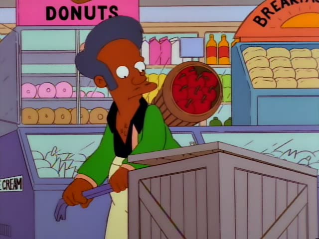 Apu works behind a wooden crate in the store, with a basket of red apples on the counter behind him