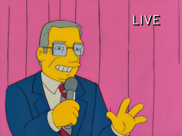 A grey-haired man with glasses, a blue suit, and a red tie holds a microphone and hosts a live broadcast on a pink-curtained…