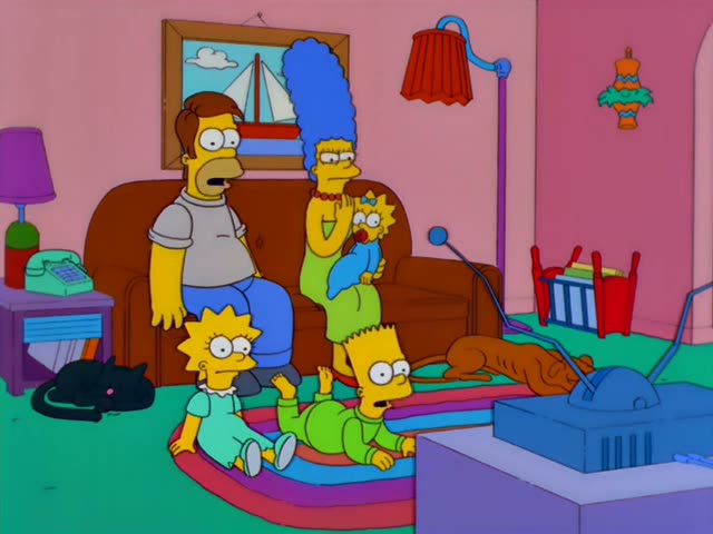 Marge stands on the couch holding baby Maggie with her pacifier and blue bow, while a heavy man with brown hair in a grey…