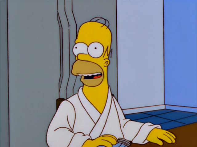 Homer Simpson sits at a wooden counter with blue tiles behind him and a basket of muffins tied with a purple bow