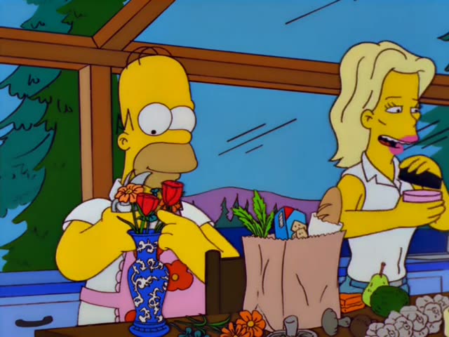 Homer arranges flowers in a blue vase in a kitchen with pine trees visible through the high windows, while a blonde woman…