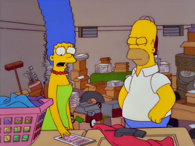 Marge and Homer stand in a cluttered storage room filled with cardboard boxes, stacks of paper, a pink laundry basket…