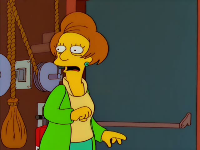 A woman with a tall auburn bouffant and a green cardigan over a yellow top stands beside a grey door with a long push bar…