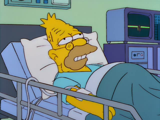 Grampa lies in a hospital bed with a drip line attached, a blue gown and green blanket over him, while a green-screen…