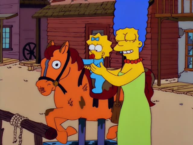 Marge stands beside a wooden rocking horse in a Western-style town while Maggie sits on the horse and reaches up to its head