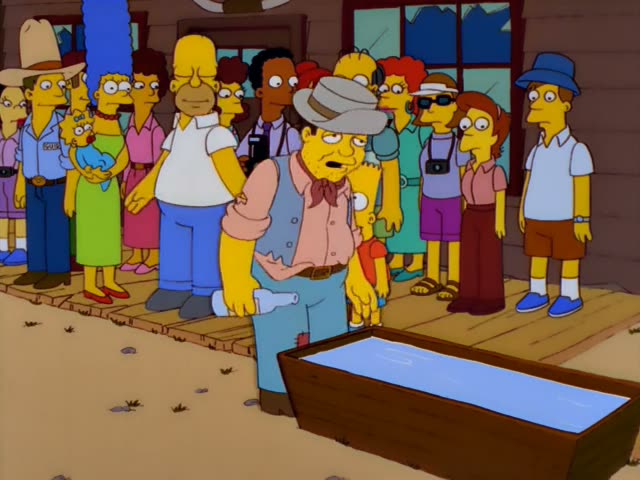 Homer, Marge with her tall blue beehive, Lisa in her red dress, and Bart in his orange shirt stand with a crowd of townsfolk…