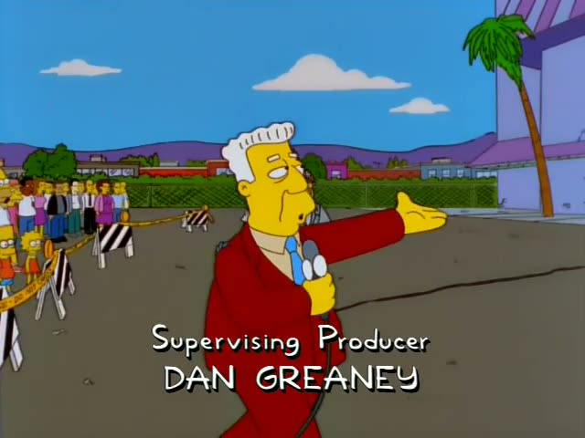 Supervising Producer Dan Greaney appears in front of a crowd of onlookers behind police tape, with a workman in a hard hat…