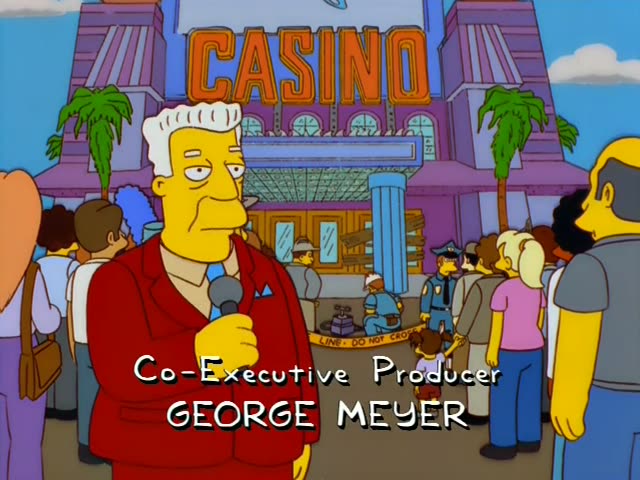 Kent Brockman stands outside the Monty Burns Casino holding a microphone and speaking to a crowd, while a bald man in grey…