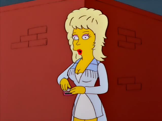 A woman with a big teased blonde hairdo, red lipstick, and a light blue dress stands against a red brick corner…