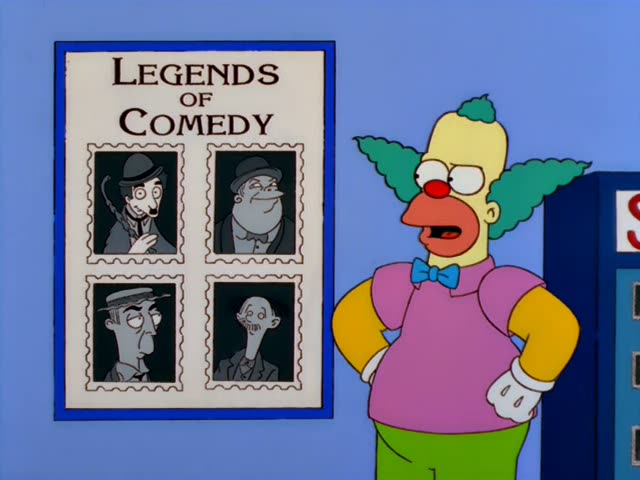 Krusty the Clown stands beside a blue wall poster titled 'Legends of Comedy' that shows four grey portraits of men in hats…