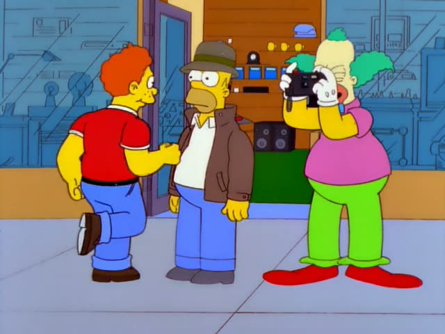 Krusty the Clown stands on a sidewalk outside a storefront raising a black device to his face, while Homer in a brown…