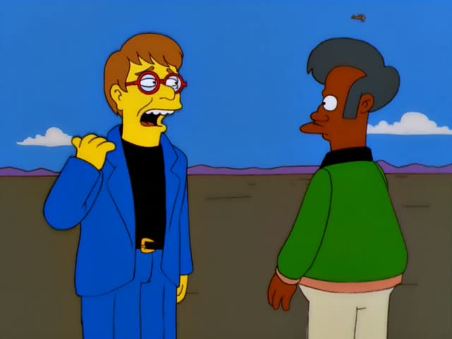 A man in a blue suit with red-rimmed glasses and a black turtleneck stands beside a dark-skinned man with a grey pompadour…