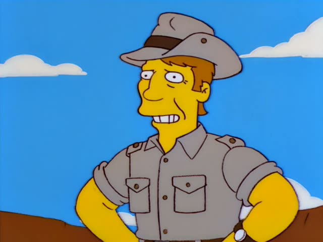 A man in a grey safari hat and grey shirt with rolled sleeves, who is not identifiable as a regular, stands against a blue…