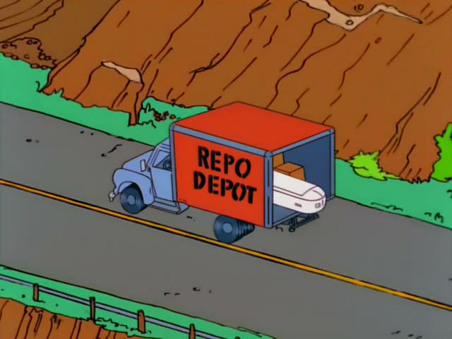 A bald man in a grey Repo Depot shirt sits in the passenger seat of a blue-and-red Repo Depot truck, dangling a small…