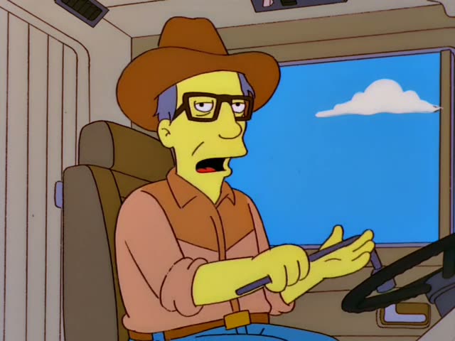 A man in a cowboy hat and dark-framed glasses sits behind a steering wheel in a cab, gesturing with one hand…