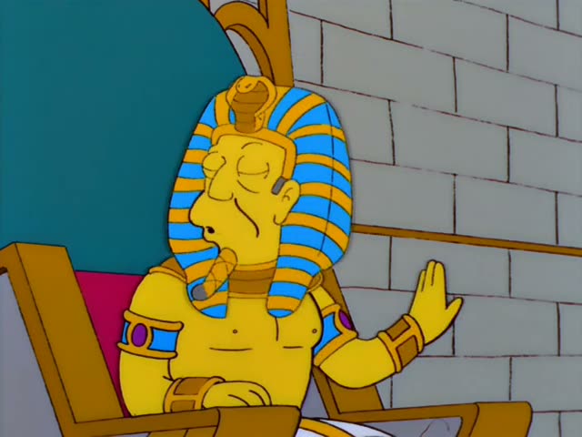 A golden-skinned Pharaoh with a striped blue-and-gold headdress and cobra ornament, who is Mr. Burns, sits on a throne…